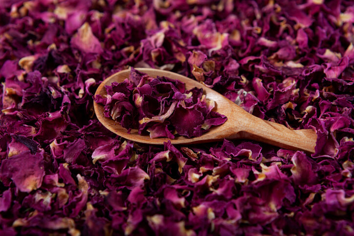 A wooden spoon resting on a bed of dried rose petals, which are deep purple in color, covering the entire surface beneath.