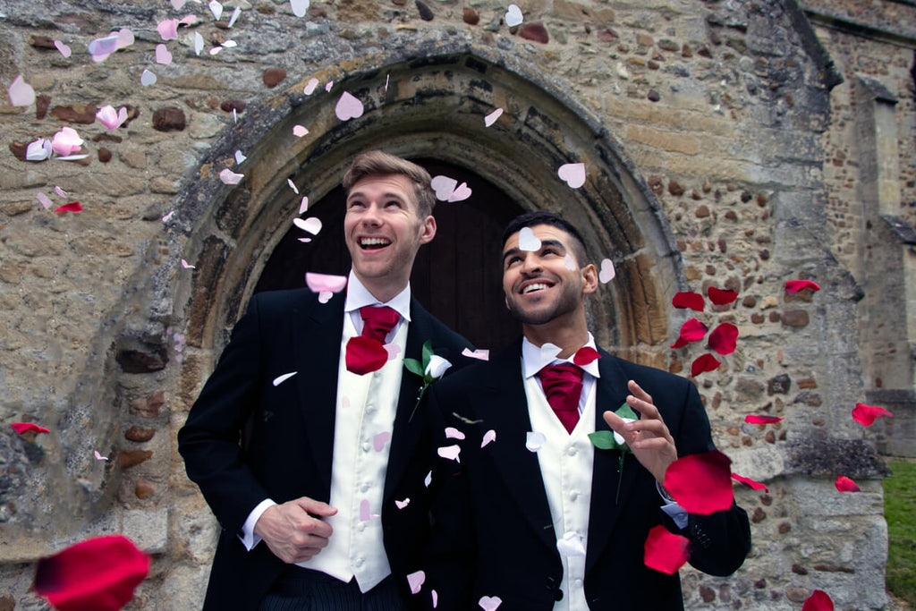 Two grooms in black suits with white shirts and red ties standing in front of an old stone church doorway, smiling joyfully as pink and white flower petals are thrown over them, celebrating their wedding day.
