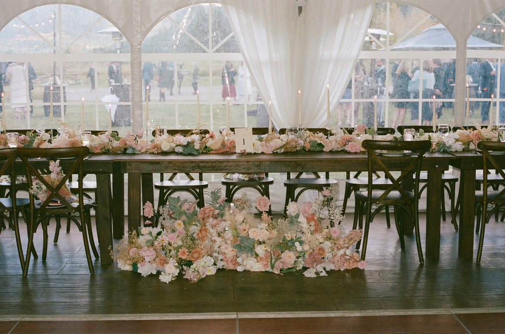 Lush floral arrangements by Hidden Botanics in soft pink and white hues adorn a long banquet table in a tented reception area, with guests mingling in the background.