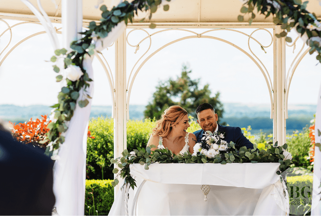 Bride and groom sharing a joyous moment at their wedding ceremony, seated behind a floral arrangement on a table under a white gazebo, with a scenic outdoor backdrop.