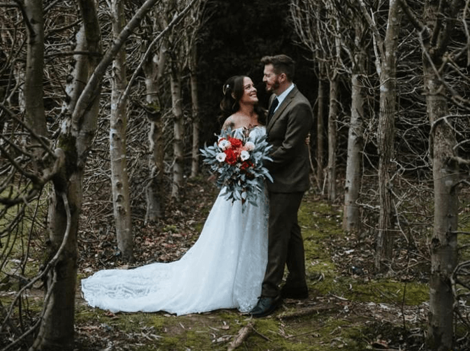 Wedding couple embracing under a canopy of trees, holding a cascading bridal bouquet in a serene woodland setting.