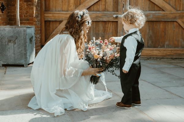 A bride holding a bouquet of artificial flowers from Hidden Botanics outside with a young boy gently touching it.
